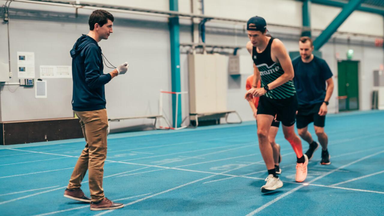 Athletes running on an indoor track while a coach times them.