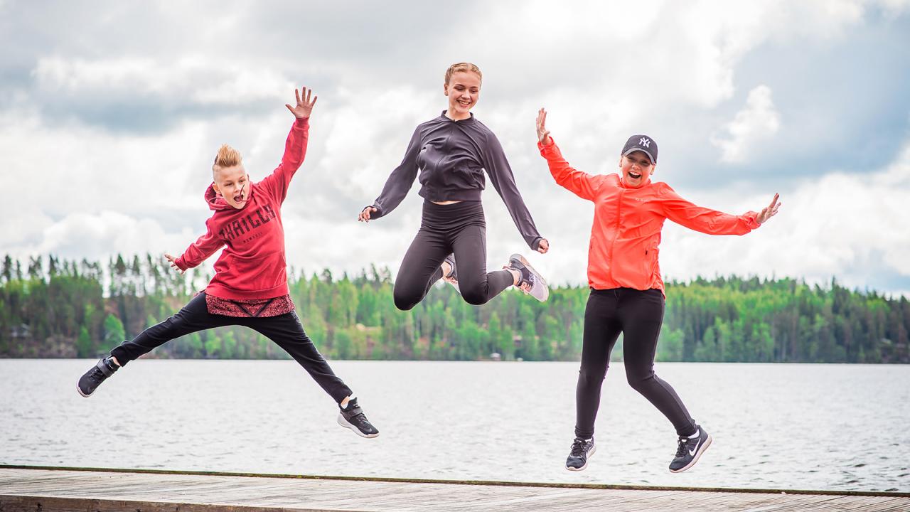 Kolme ihmistä hyppää laiturilla järven edessä, taustalla metsää.