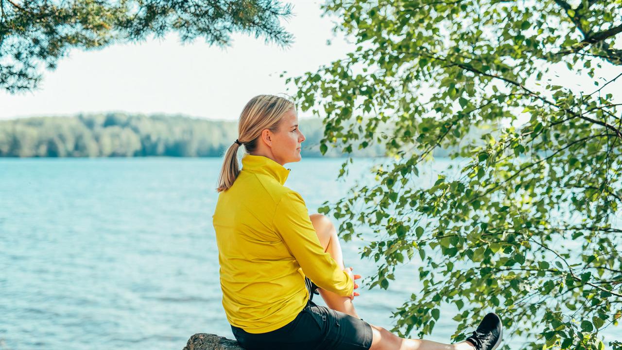 Nainen istuu kalliolla järven rannalla, taustalla puita.