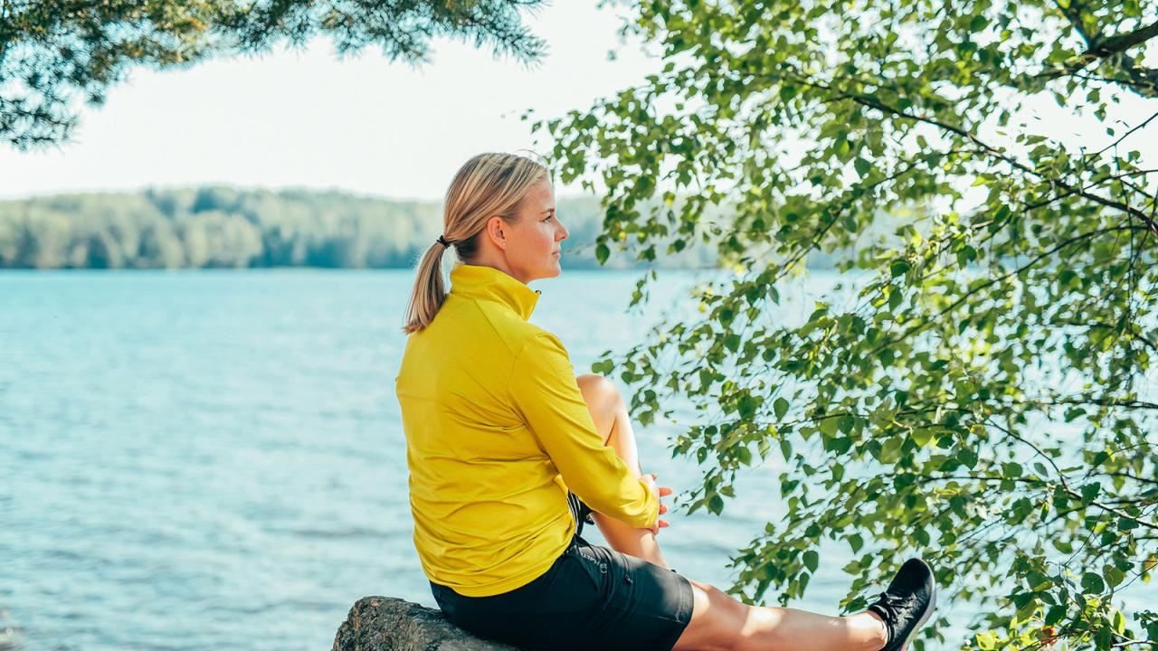 Nainen istuu kalliolla järven rannalla, taustalla puita.