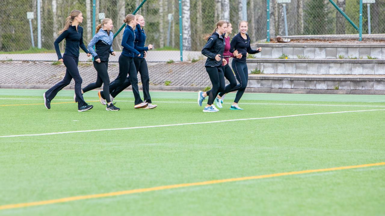 Nuoria juoksemassa urheilukentällä, taustalla puita ja katsomo.