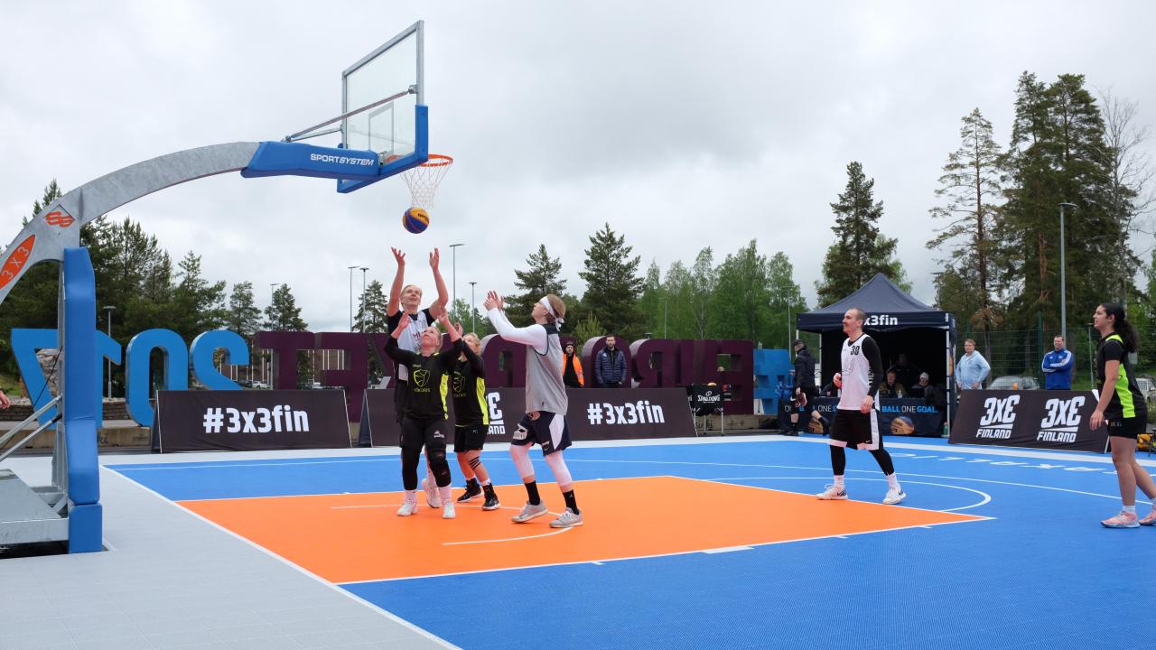 Ulkokoripallokentällä pelataan 3x3-ottelua. Pelaajat hyppäävät palloa tavoitellen.