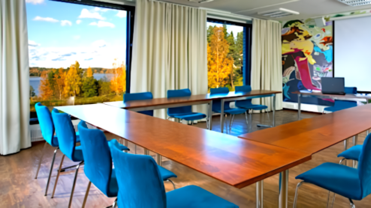 Kokoushuoneessa u-muotoinen pöytä, siniset tuolit, taulu ja ikkunat syksyiseen maisemaan.