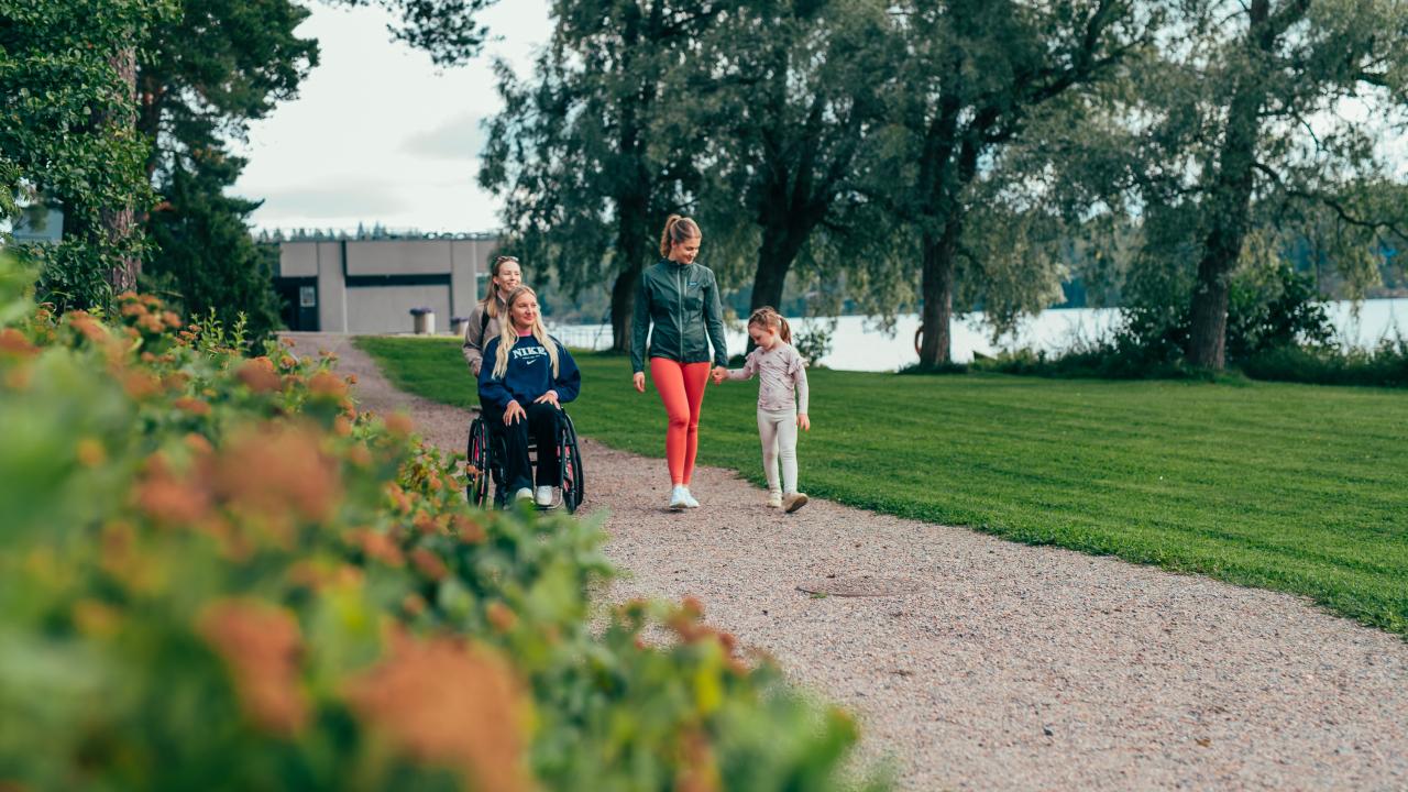 Nainen työntää pyörätuolissa nuorta naista, ja vierellä kävelee toinen nainen pidellen kädestä pientä tyttöä.