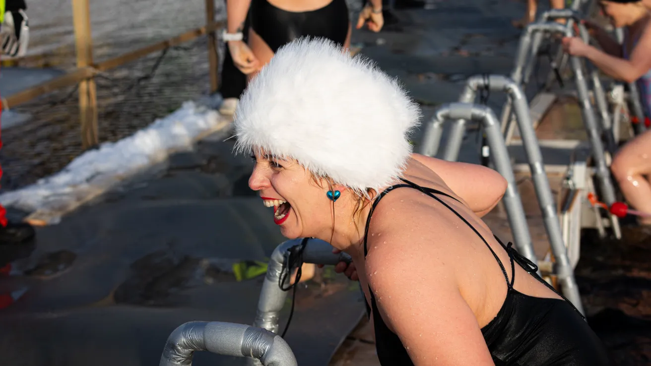 Naurava nainen jäisessä vedessä, pukeutunut mustaan uimapukuun ja valkoiseen hattuun.