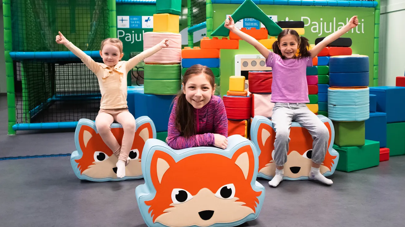 Kolme lasta leikkipaikalla, hymyillen ja istuen kettuaiheisten tyynyjen päällä.