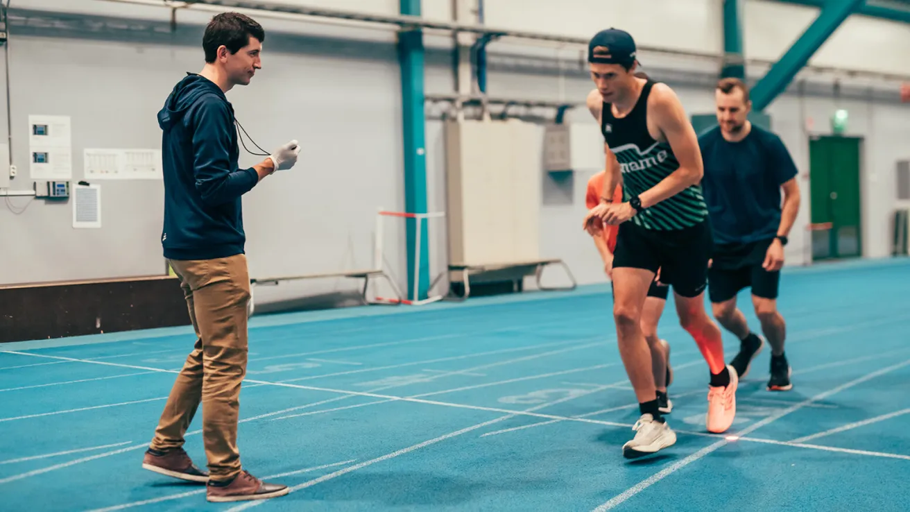 Kilpajuoksijat treenaavat sisäradalla, valmentaja seuraa.