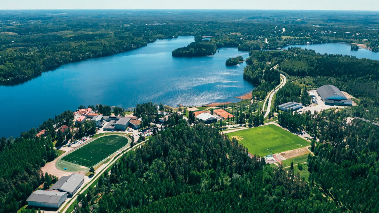 Ilmakuva järvestä ja metsäisestä maisemasta, jossa urheilukenttiä ja rakennuksia.