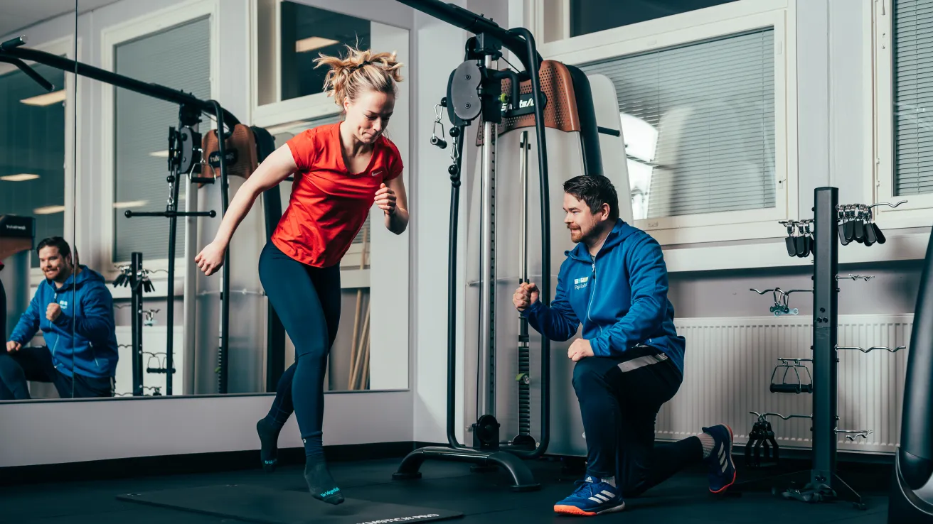 Nainen treenaa salilla, valmentaja kannustaa vieressä.