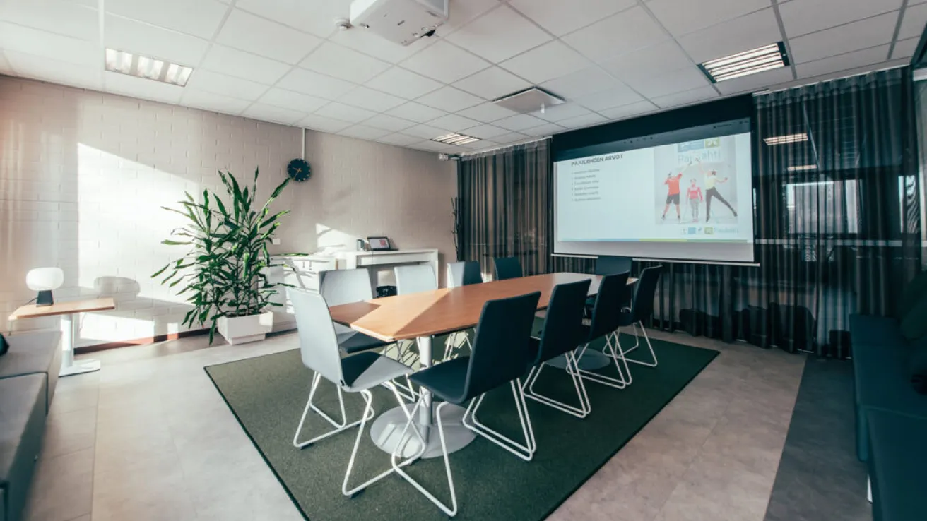 Moderni kokoushuone, valkoinen pöytä, mustat tuolit, suuri näyttö seinällä.