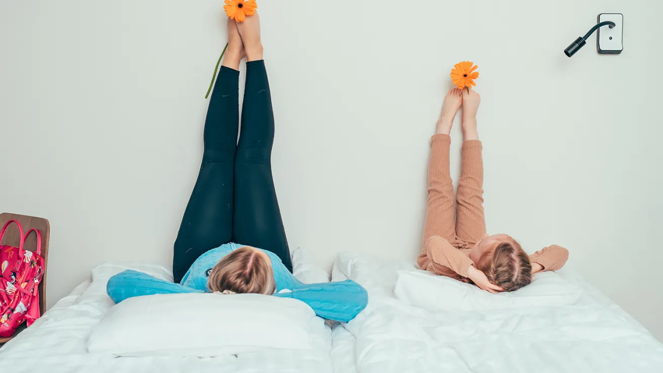 Kaksi henkilöä makaa sängyllä, jalat nostettuina seinää vasten, oranssi kukka jaloissaan.
