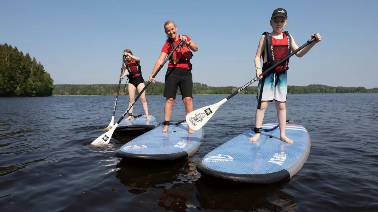 Kolme ihmistä suppailee järvellä aurinkoisena päivänä.