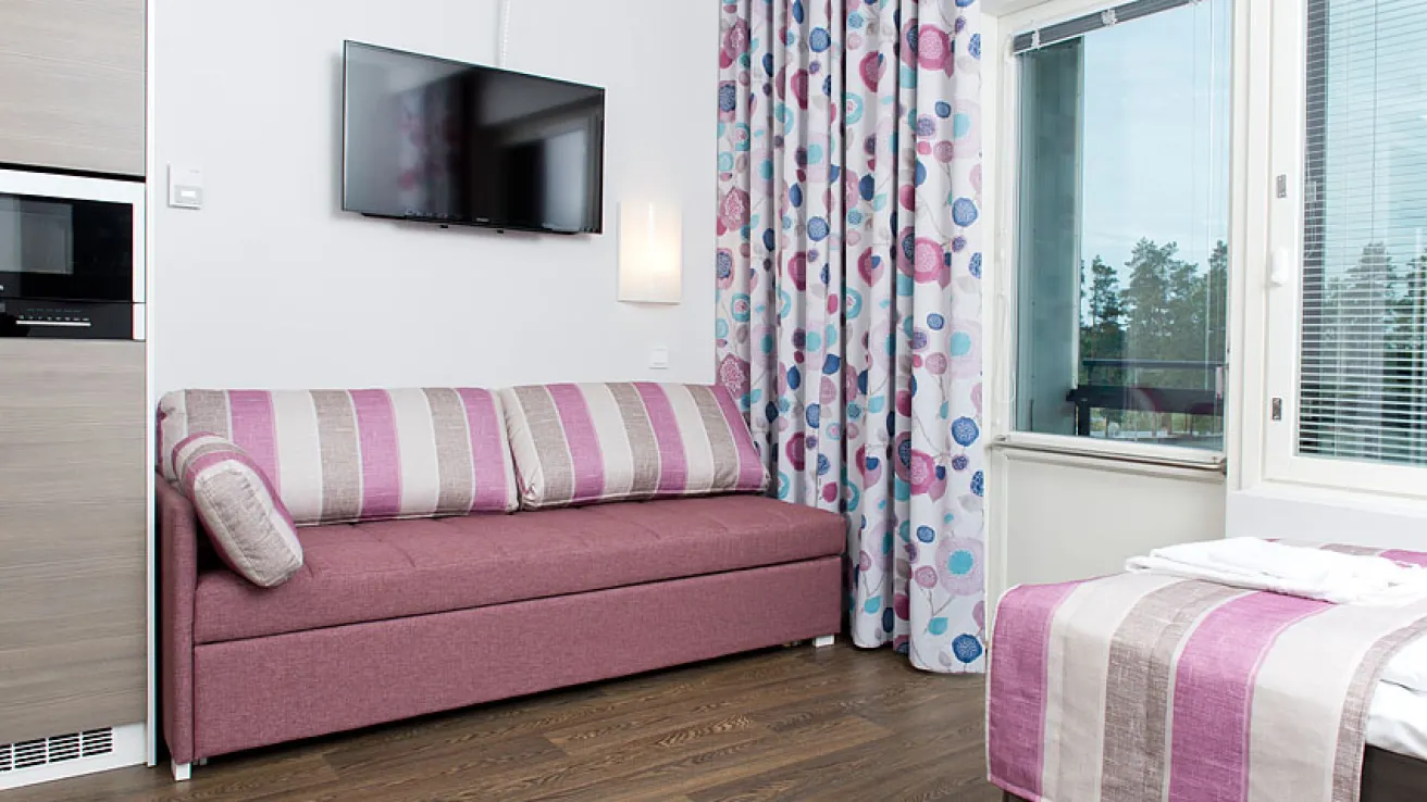 Huone pinkillä sohvalla, raidallinen sänky, televisio, värikkäät verhot.