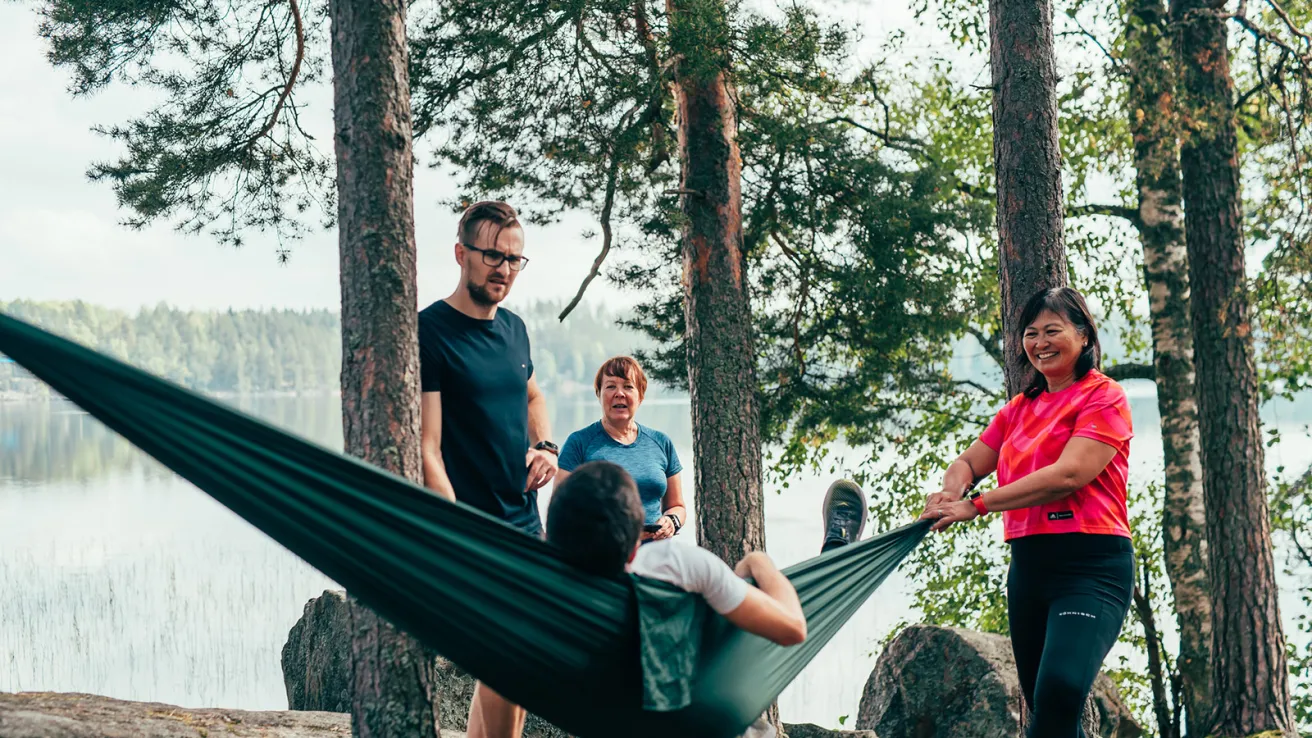 Neljä ihmistä puiden keskellä järven rannalla, yksi makaa riippumatossa.