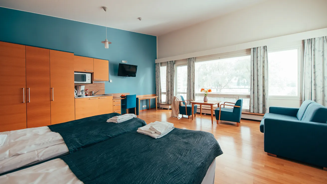 Hotellihuone, iso ikkuna, siniset ja puiset sävyt, leveä sänky ja työpöytä.