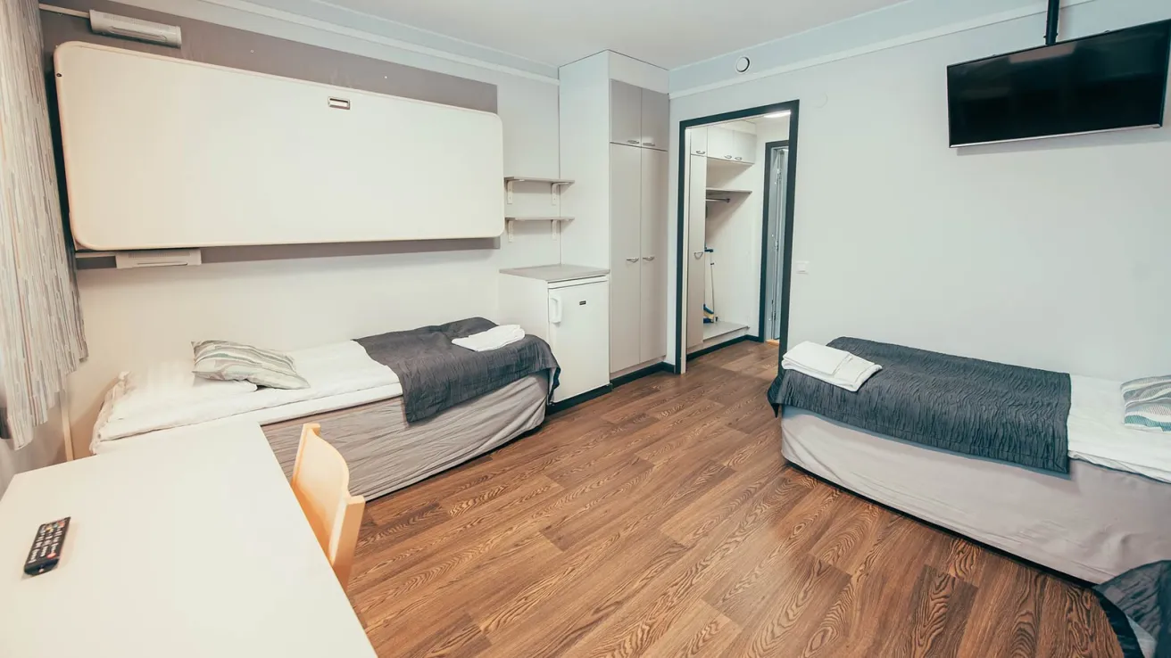 Kaksi sänkyä ja televisio vaaleassa makuuhuoneessa, puulattia.