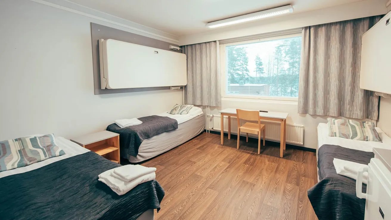 Kaksi sänkyä huoneessa, ikkuna ja pöytä, vaalea sisustus.