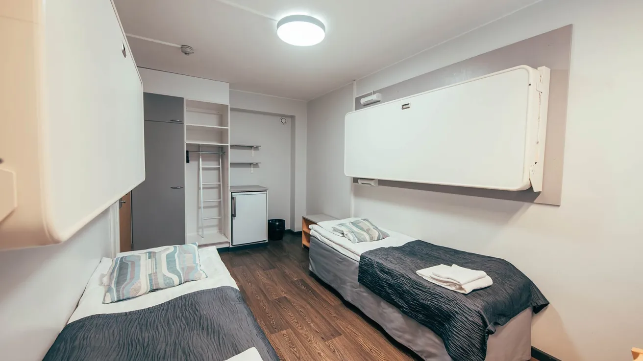 Kaksi sänkyä huoneessa, vaalea seinä ja puulattia.