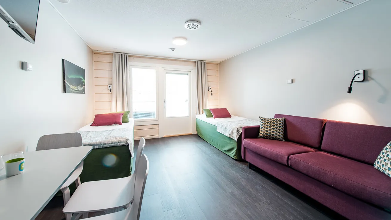 Valoisa hotellihuone, kaksi sänkyä, sohvapöytä, violetti sohva.