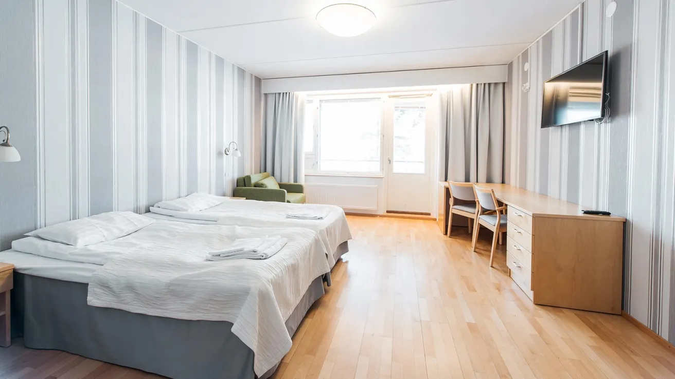 Valoisa hotellihuone, jossa on kaksi sänkyä, pöytä ja televisio.