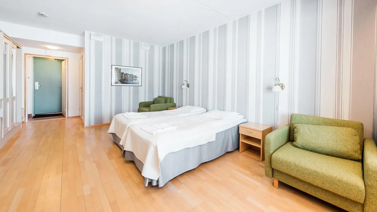 Valoisa hotellihuone, kaksi sänkyä ja vaaleanvihreät nojatuolit.