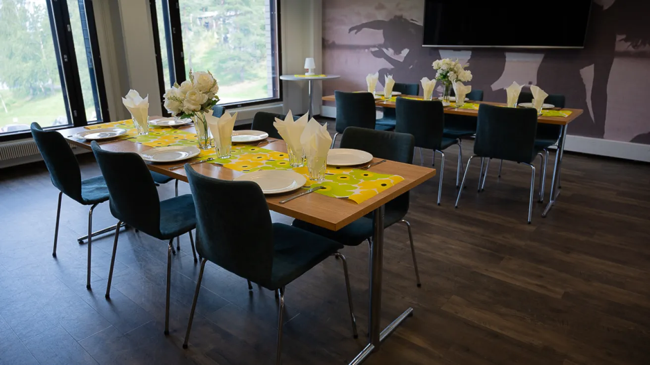 Kahden pitkän pöydän kattaus valkoisin lautasin ja kukkakoristein huoneessa.