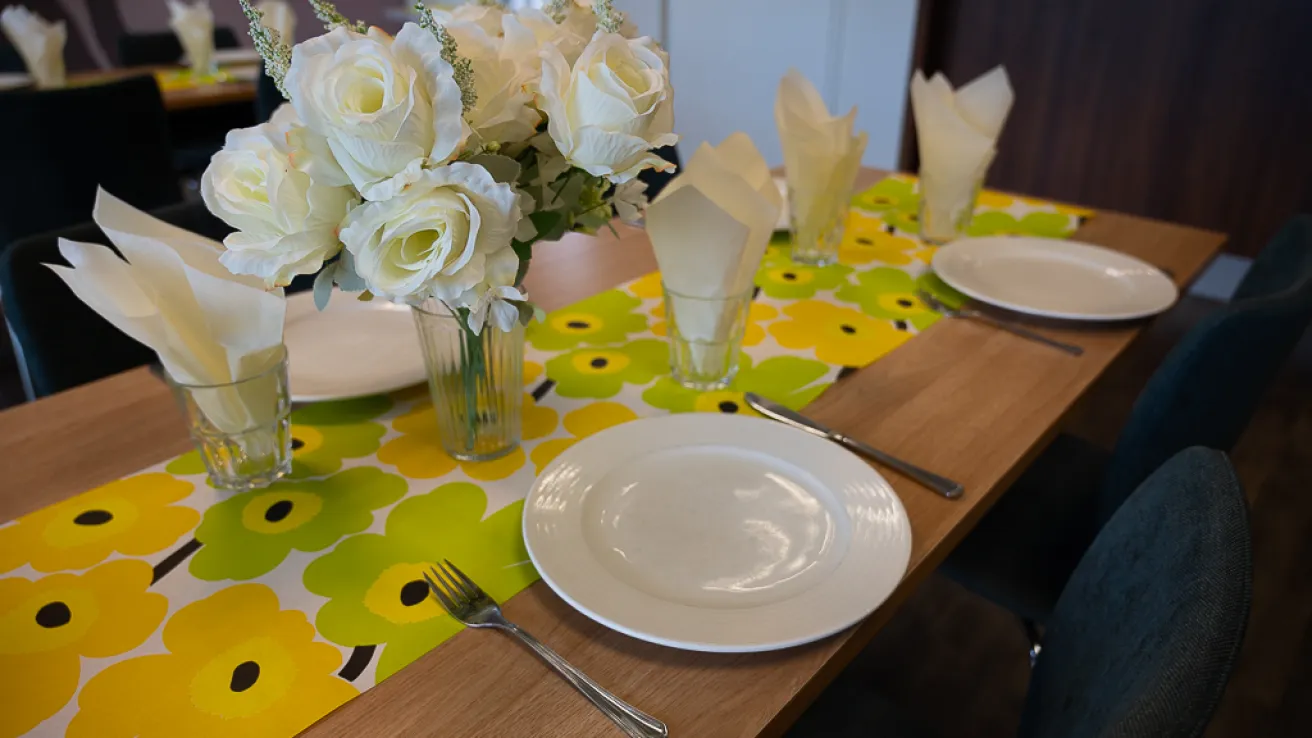 Pöytä vihreäkukkaliinalla, valkoisia lautasia ja kukkakimppu.