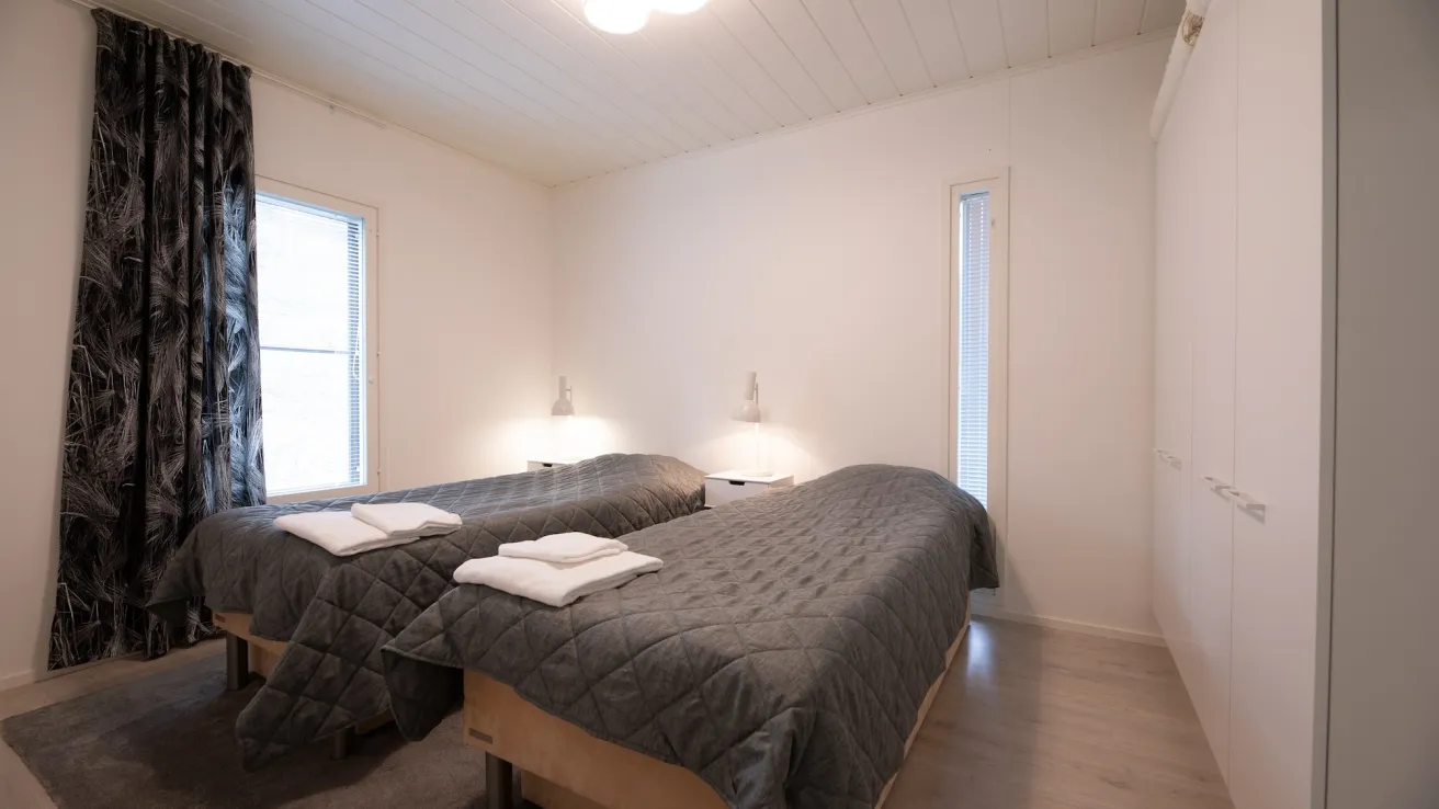 Kaksi sänkyä valkoisessa makuuhuoneessa, harmaat peitot, ikkunat vasemmalla.