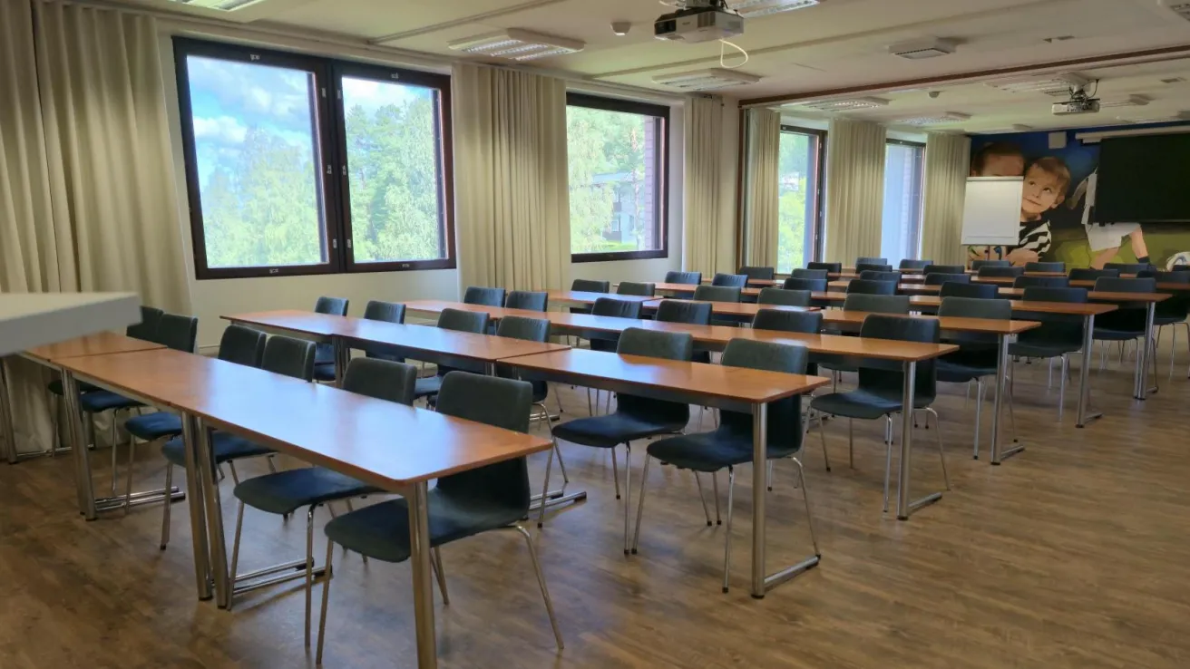 Tyhjä luokkahuone, puiset pöydät, mustat tuolit, ikkunat taustalla.