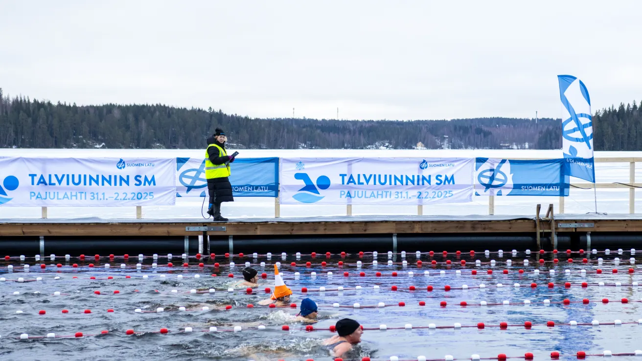 Talviuimarit kilpailemassa ulkoaltaassa, taustalla metsää ja kyltti.