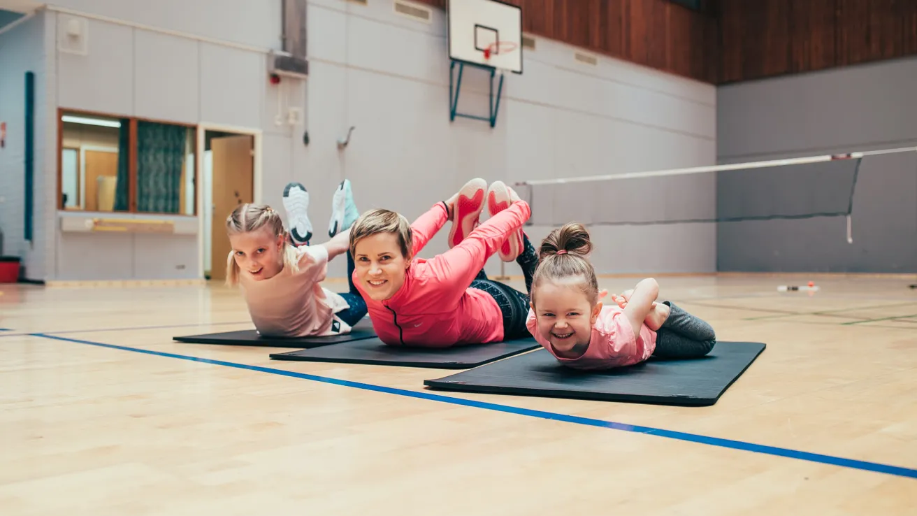 Kolme lasta jumppasalissa tekemässä selkärullia kuntomatoilla.
