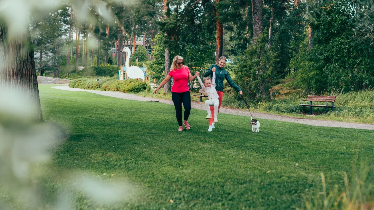 Kaksi ihmistä kävelee nurmikolla koiran kanssa, puisto taustalla.
