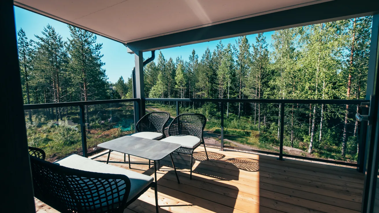 Puistopaju. Puinen terassi metsämaisemalla, pöytä ja tuolit varjossa.