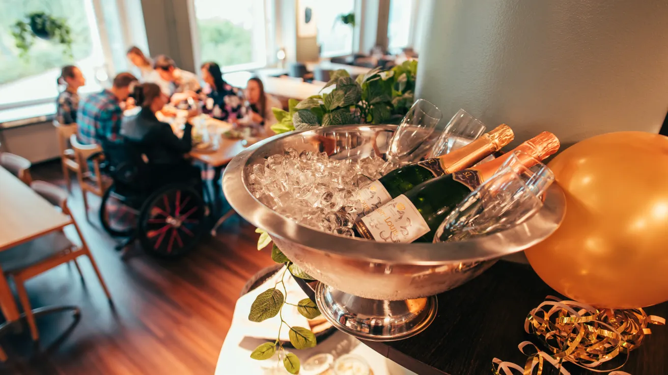 Kuohuviini jääpala-astiassa vappupallojen vieressä seurueen ruokaillessa taustalla.