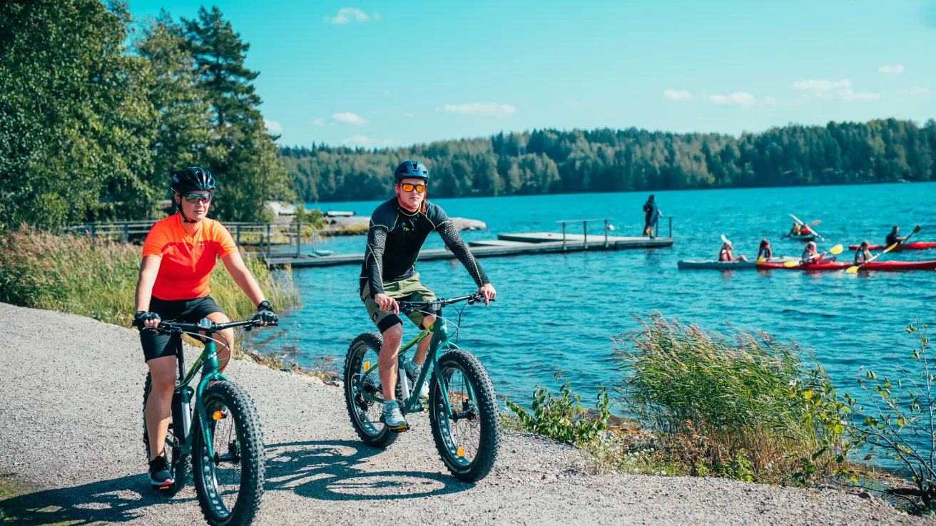 Mies ja nainen pyöräilevät fatbikeillä järven rannalla ja taustalla ihmisiä melomassa.
