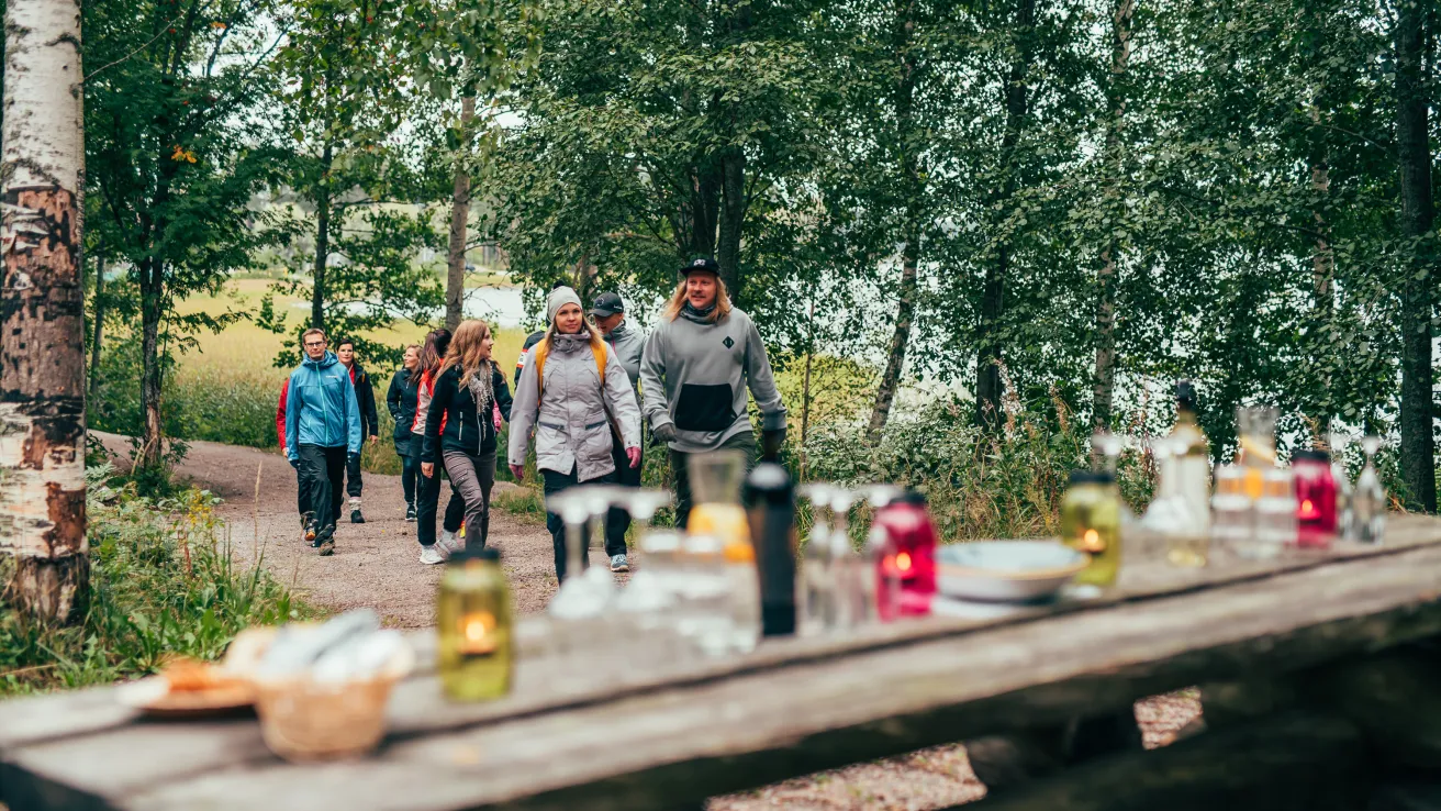 Polulla kävelee useita ihmisiä metsässä. Etualalla on katettu puupöytä tuikkuineen..