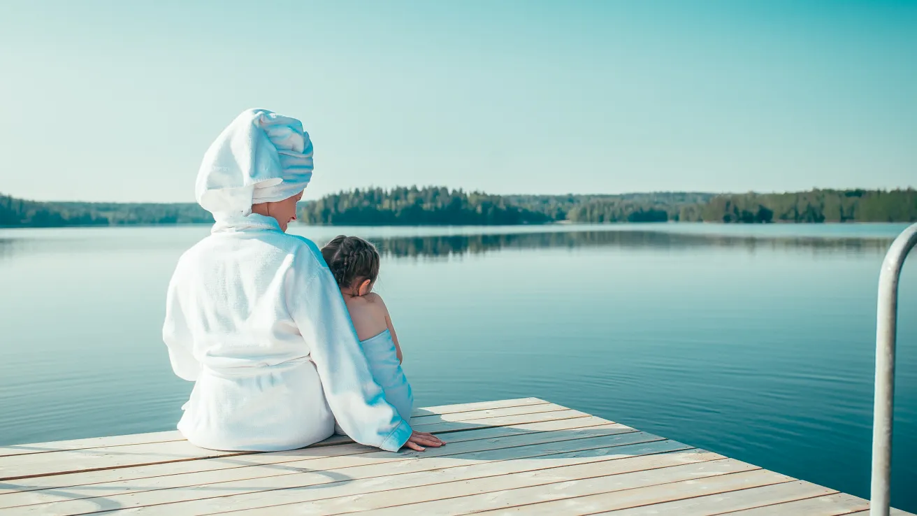 Nainen valkoisessa kylpytakissa ja tyttö pyyhe ympärillään istuvat laiturilla katsellen tyyntä, kesäistä järveä.