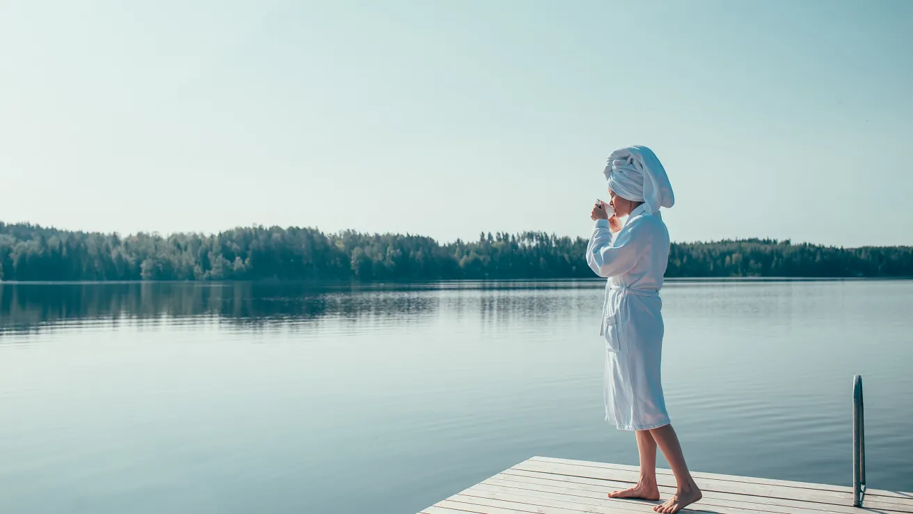 Nainen kylpytakissa siemailee kahvikuppia Pajulahden laiturilla tyynen järven äärellä.