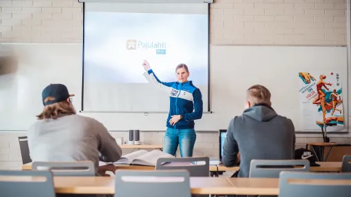 Luokkahuone, opettaja näyttää diaa, opiskelijat istuvat pöydissä.