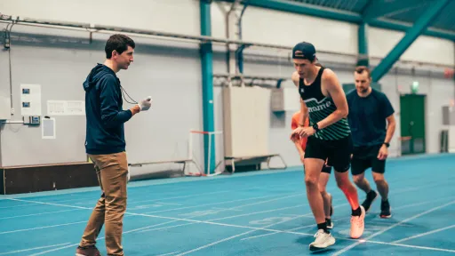 Athletes running on an indoor track while a coach times them.