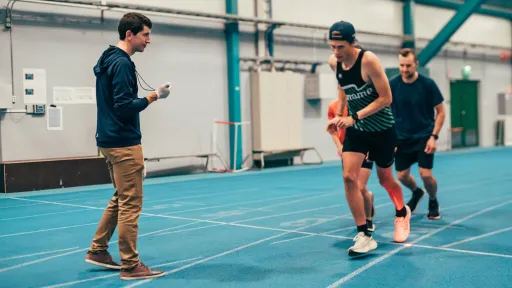 Athletes running on an indoor track while a coach times them.
