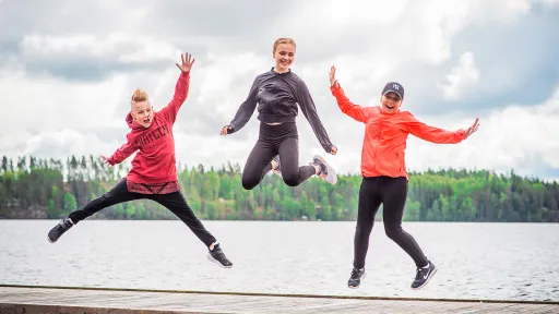 Kolme ihmistä hyppää laiturilla järven edessä, taustalla metsää.