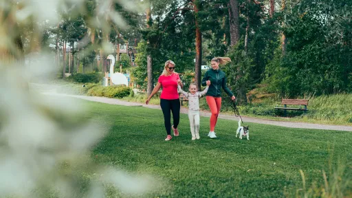 Pajulahden rantanurmella iloinen lapsi pitää kädestä mummoa ja äitiä koiran kävellessä vierellä.