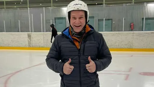 A man smiling and giving a thumbs-up on an ice rink, wearing a helmet and jacket.