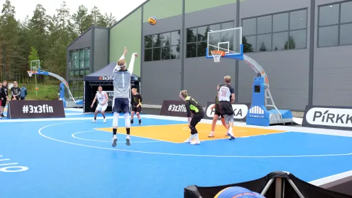Outdoor basketball game with players on a blue court next to a building.
