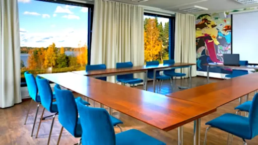 Kokoushuoneessa u-muotoinen pöytä, siniset tuolit, taulu ja ikkunat syksyiseen maisemaan.