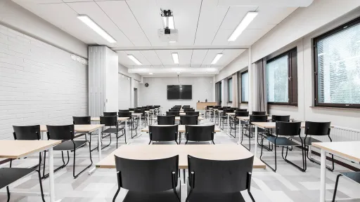 Tyhjä luokkahuone, rivissä mustia tuoleja ja pöytiä, seinillä valkoisia tiiliä ja ikkunoita.