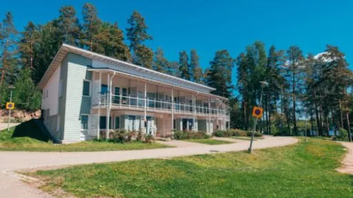 Kaksikerroksinen valkoinen rakennus vehreän metsän reunalla aurinkoisena päivänä.