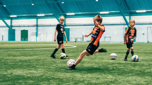 Kolme lasta pelaa jalkapalloa sisätiloissa sinisellä kentällä.