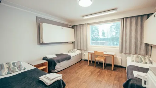 Kaksi vuoteellinen huone, jossa pöytä ja suuri ikkuna harmailla verhoilla.